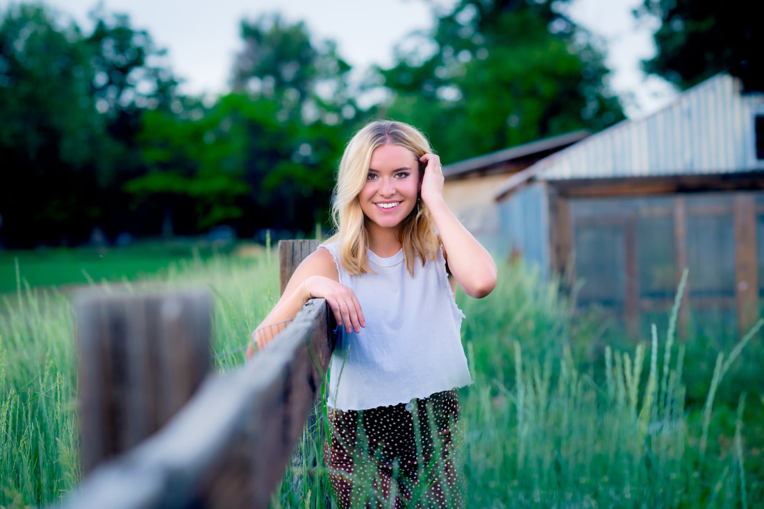 Girl wearing white shirt standing in a lush green field holding onto a fence for senior pictures 
