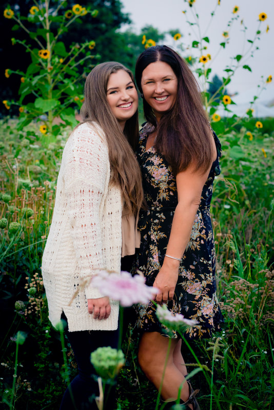 Mom in black dress, daughter in cream cardigian standing in a field of sunflowers for senior pictures