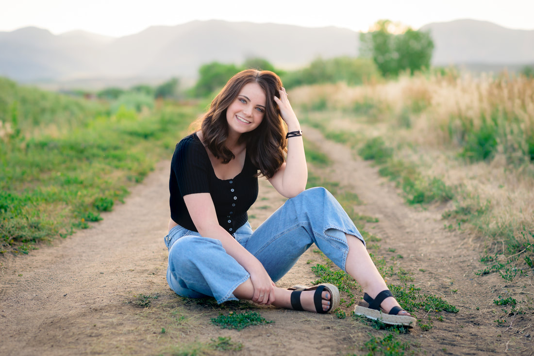 Girl in black top and jeans smiling at camera, sitting on a dirt path for senior pictures 