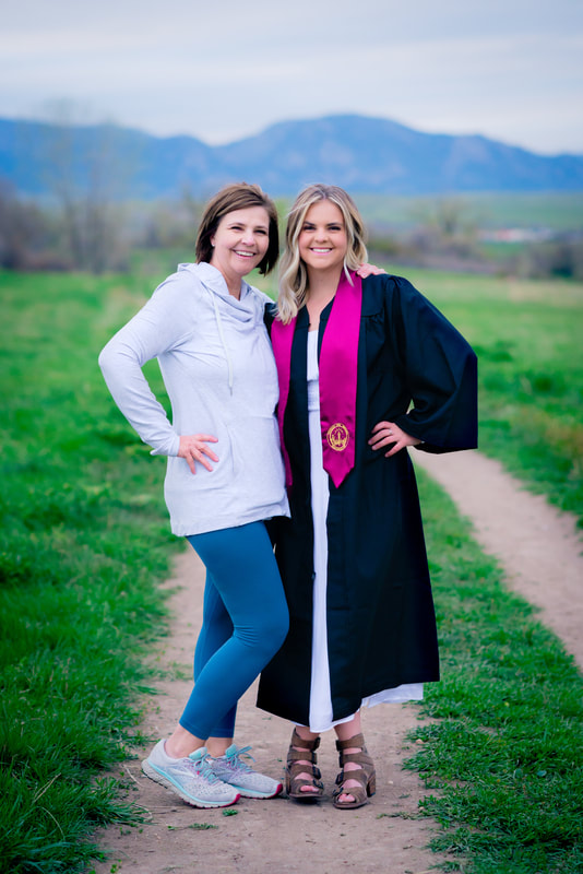 Mom in grey top, blue pants and daughter in her graduation gown standing on a dirt path together for senior pictures 