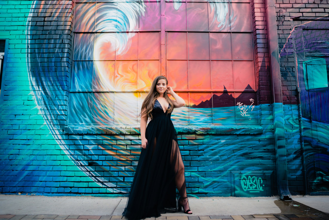A portrait of a young woman standing in front of a mural painted on bricks, touching her hair and wearing a dark floor length dress and high heels.