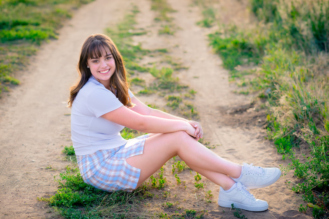 Girl with brown eyes wearing a white shirt smiling at camera sitting on a dirt path for senior pictures 
