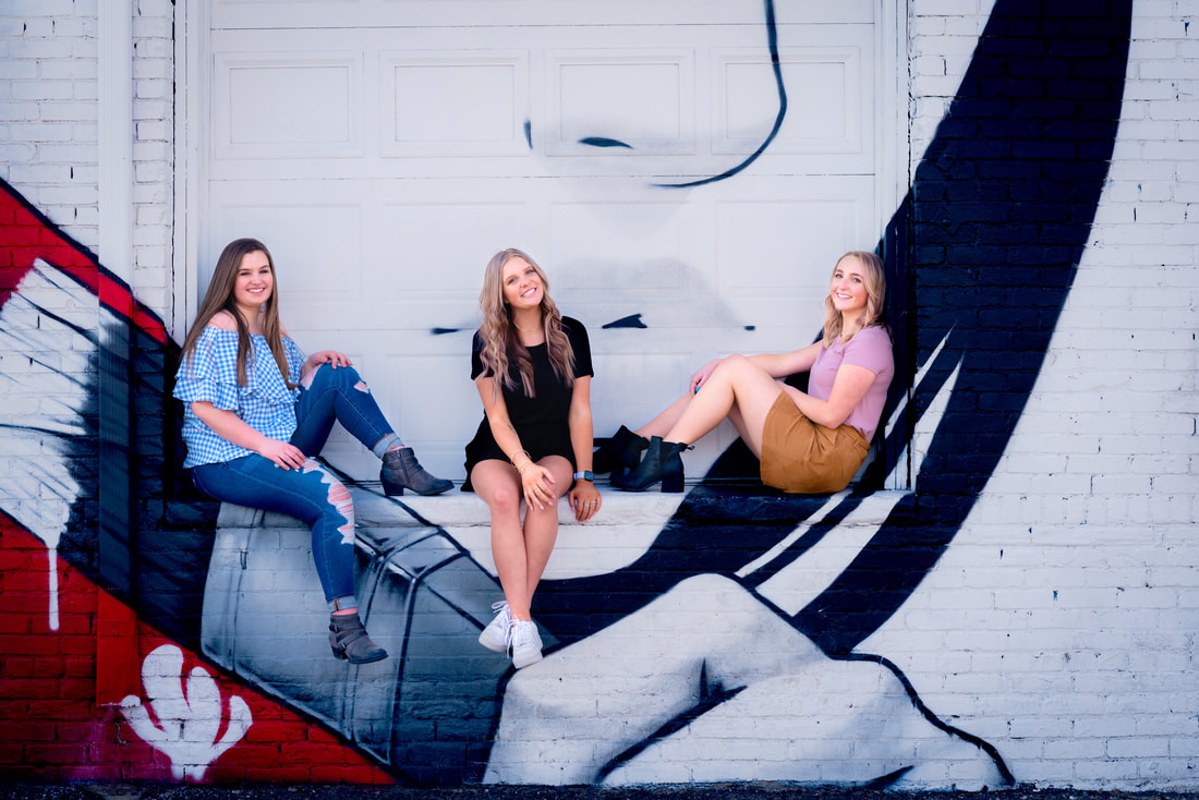 three girls sitting on brick ledge in front of colorful urban art graffiti for senior pictures