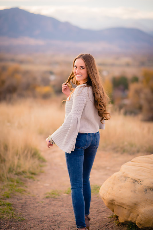 girl in grey sweater and blue jeans smiling in golden field and fall leaves with mountains behind her for senior pictures