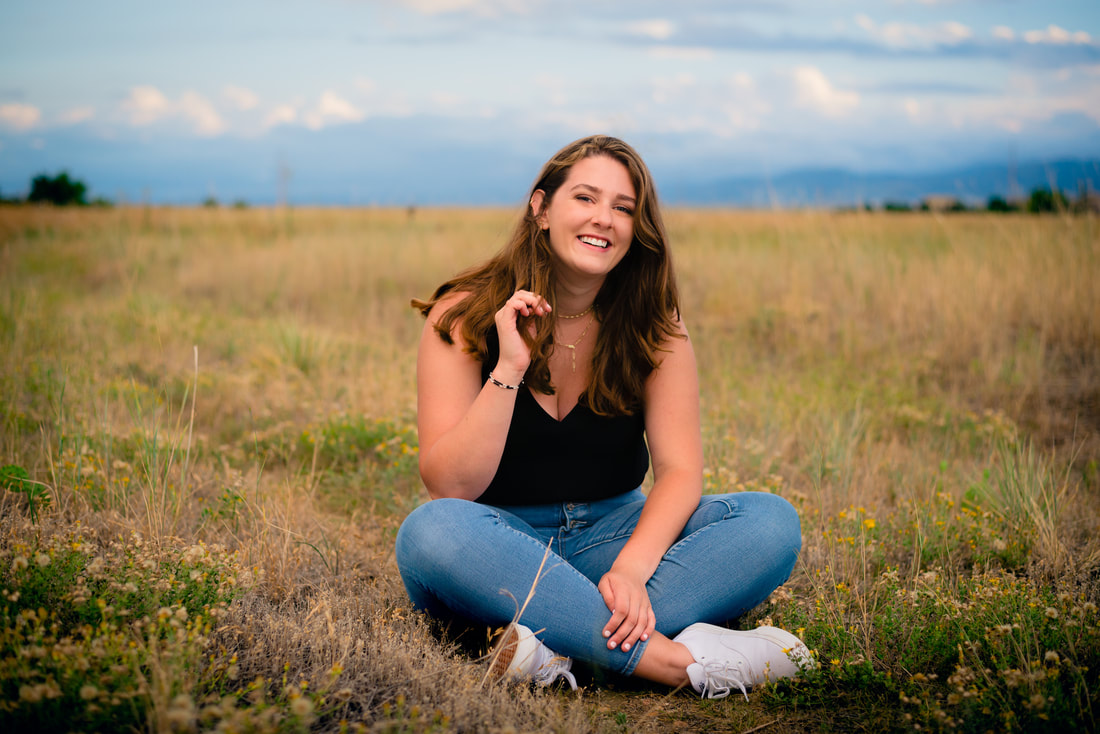 Girl in black tank top and jeans sitting in a field smiling for senior pictures