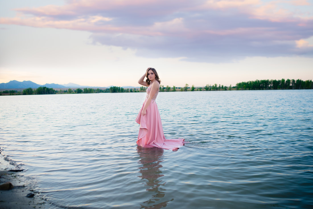 girl in pink dress standing in blue water at sunset with mountains behind her for senior pictures