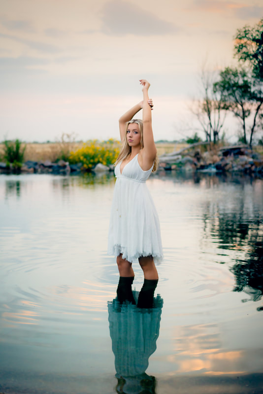 girl in white dress standing in water at sunset holding arms above her head for senior pictures